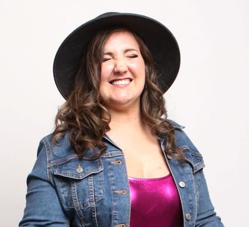 Courtney is a very happy smiling caucasian woman wearing a dark hat and a jean jacket over a shiny magenta top