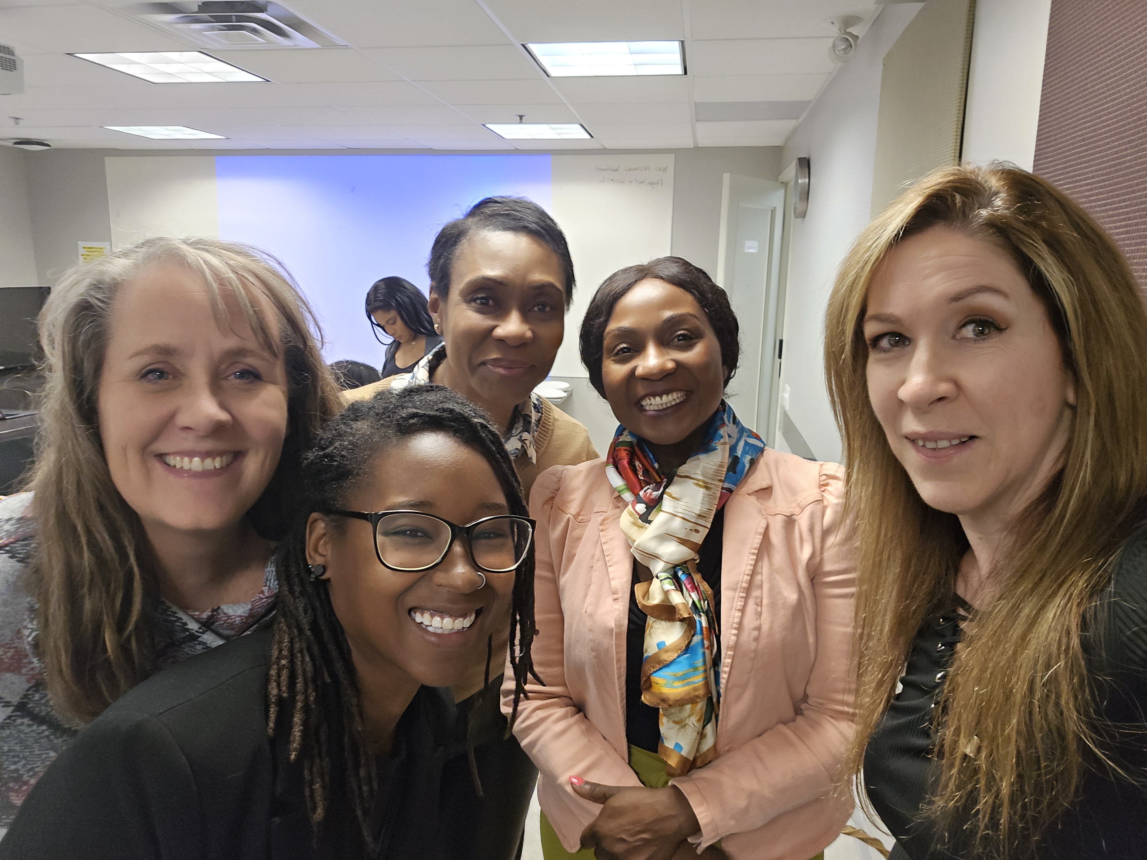 Photo of five women of various races in a meeting room smiling at the camera