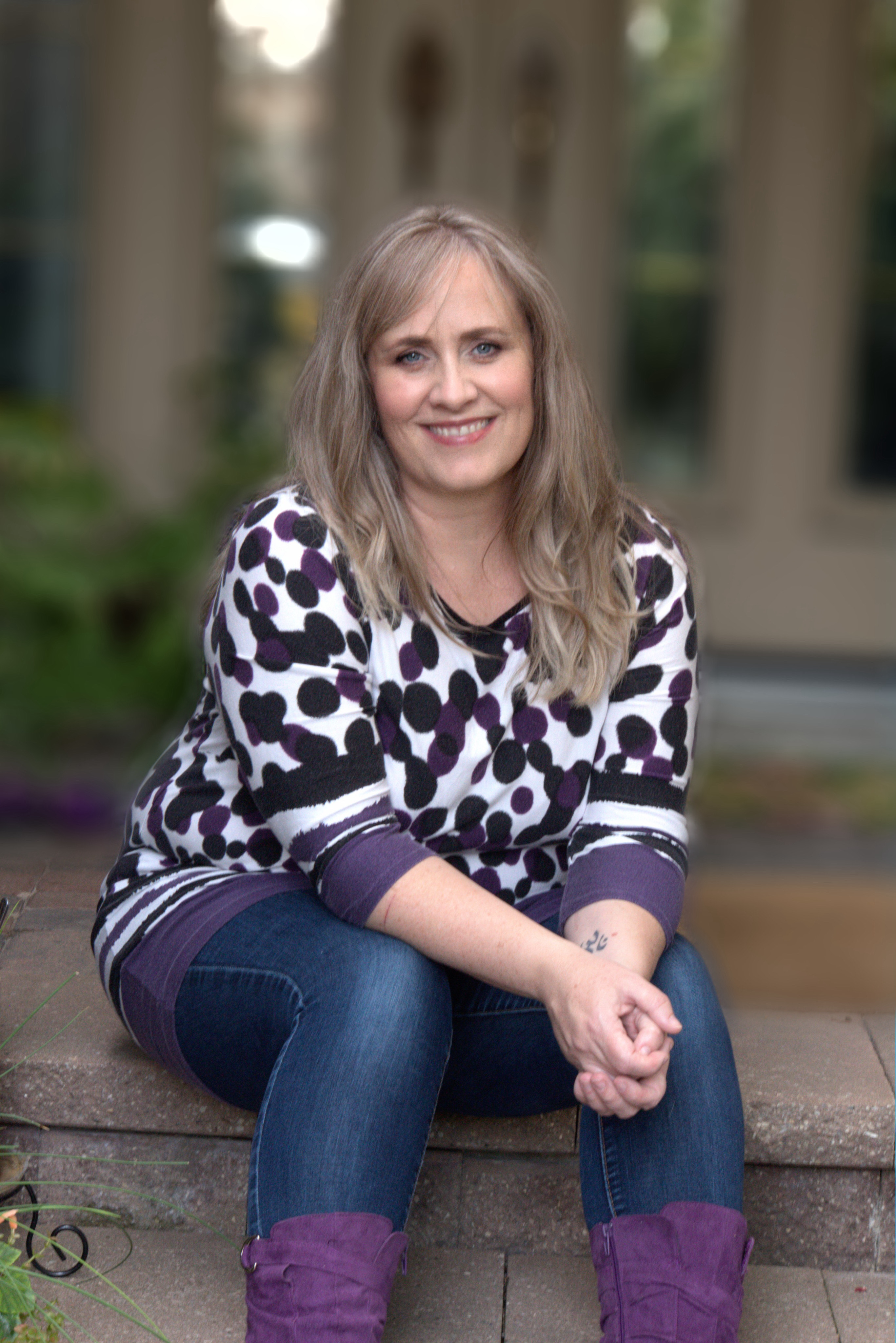 Image of a blonde woman sitting on a stone step wearning a black and purple polka dotted shirt with jeans and purple boots